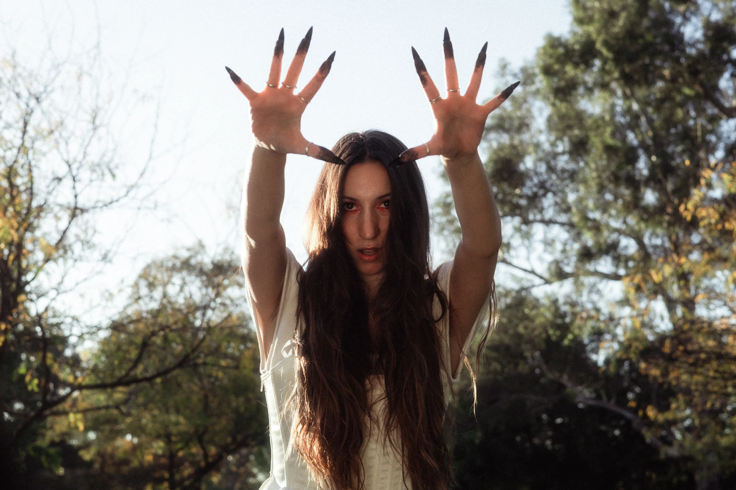 A woman with long hair extends her hands, displaying sharp, dark nails. She stands outdoors with trees and a bright sky, conveying a mystical vibe.