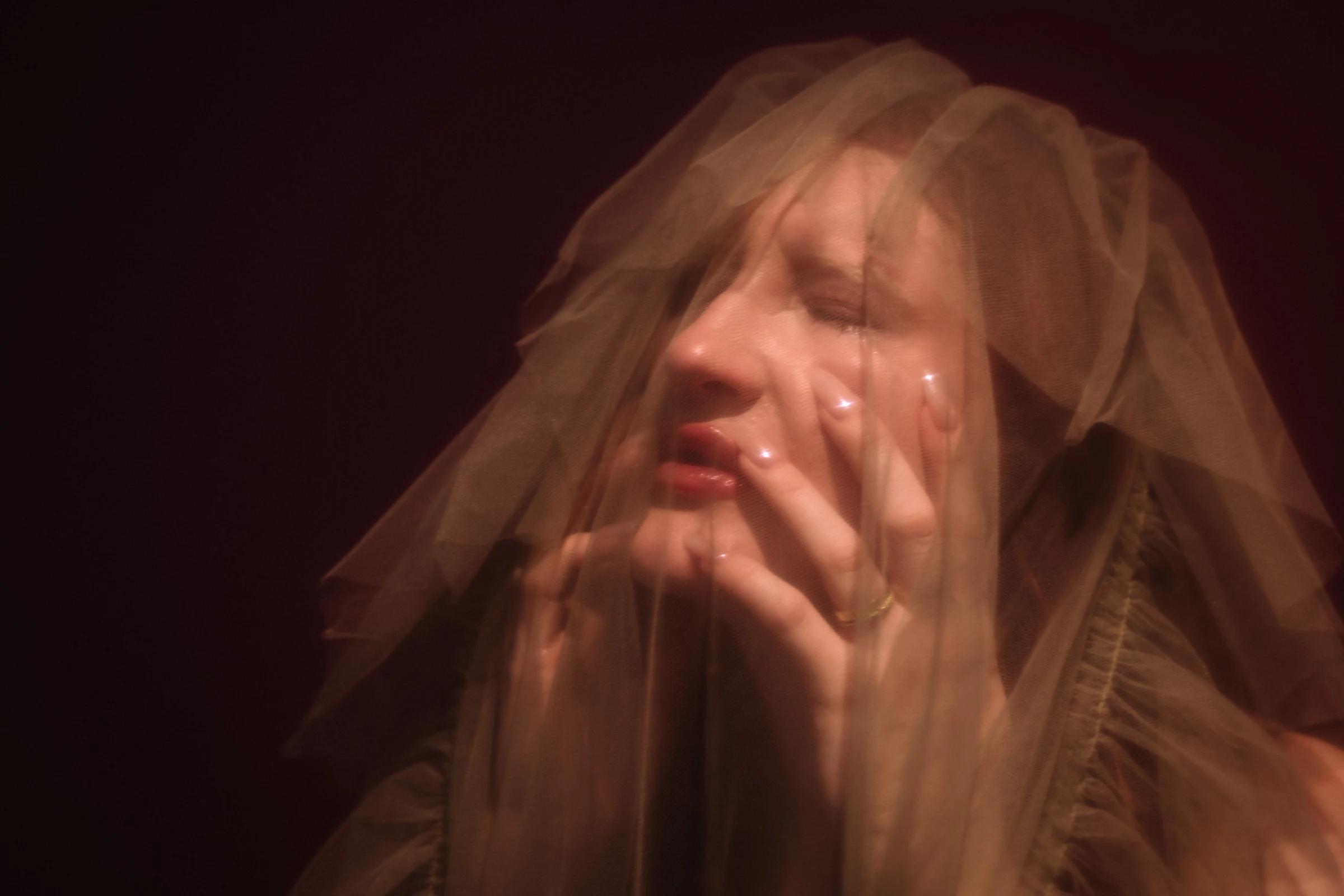 A person is covered by a translucent veil, hands touching their face, expressing emotion. The lighting is soft, casting a gentle, mysterious mood.