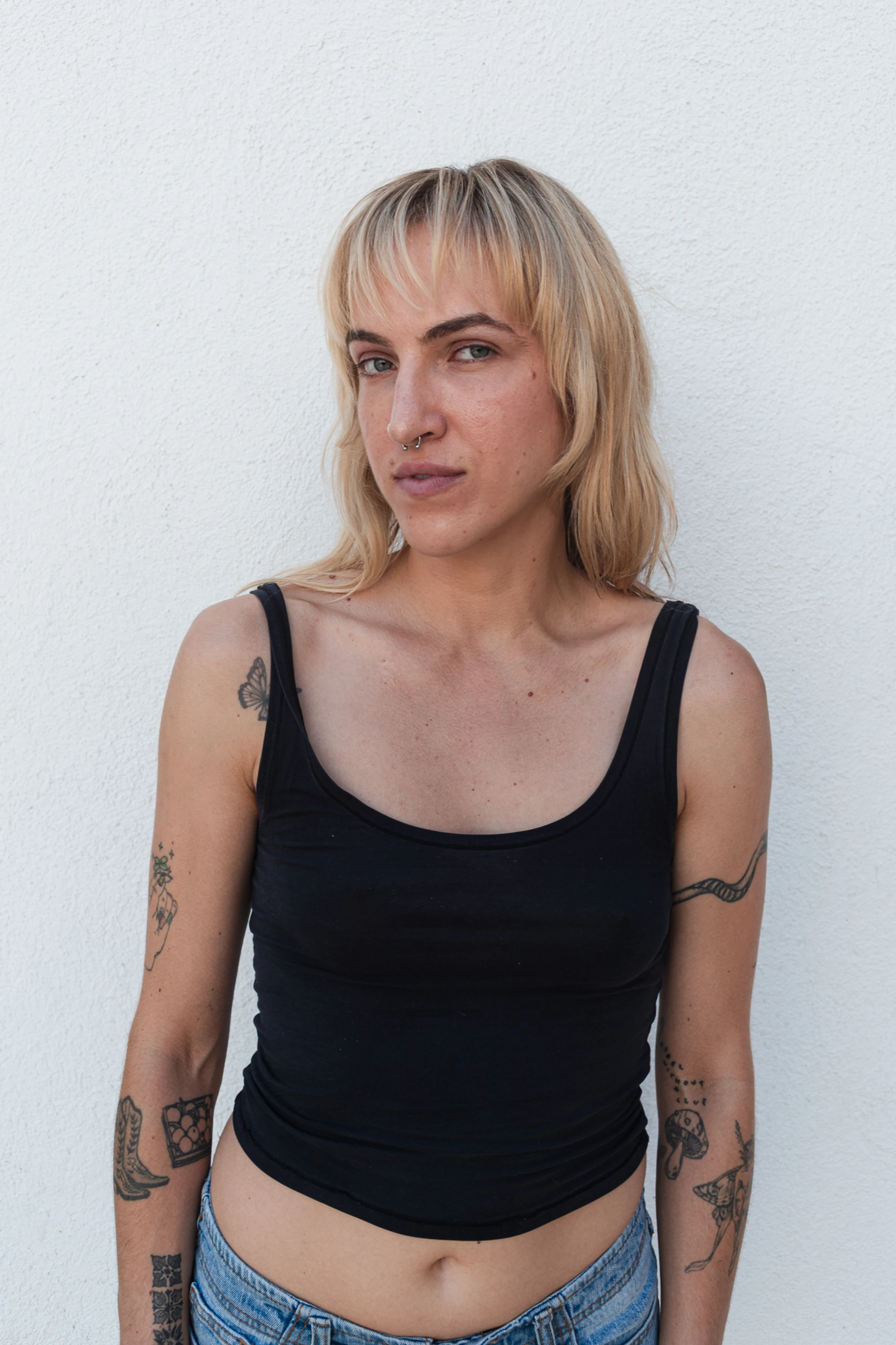 A person with medium-length blonde hair and a septum piercing stands confidently against a white background, wearing a black tank top and displaying tattoos.