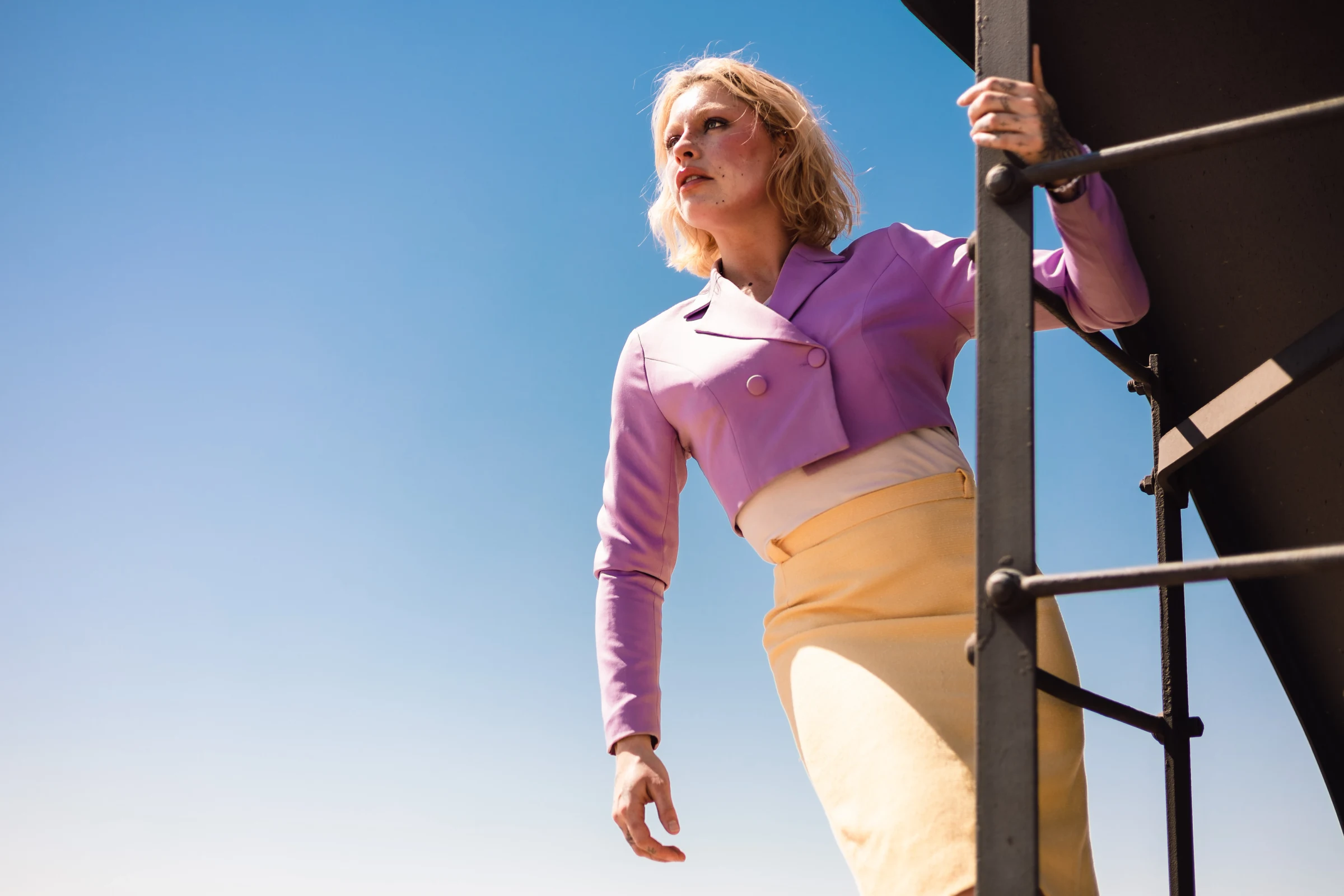 Woman in a lavender jacket and yellow skirt stands on a ladder against a clear blue sky, gazing confidently into the distance. Bright and hopeful mood.
