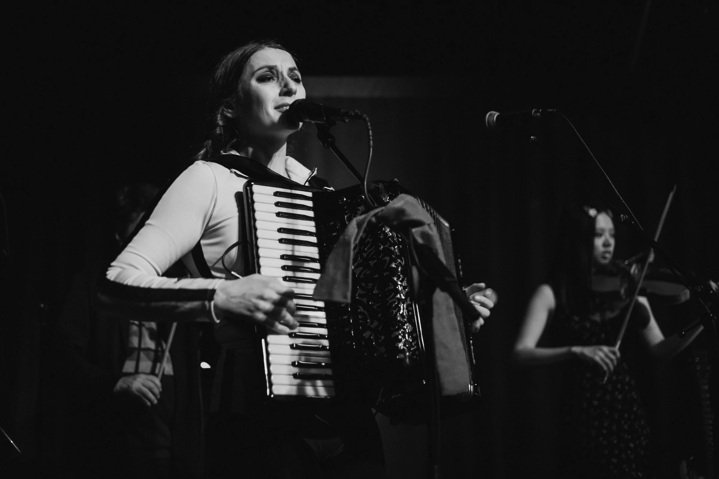 A woman passionately plays an accordion and sings into a microphone on stage, while another musician in the background plays a violin. The scene is in black and white.