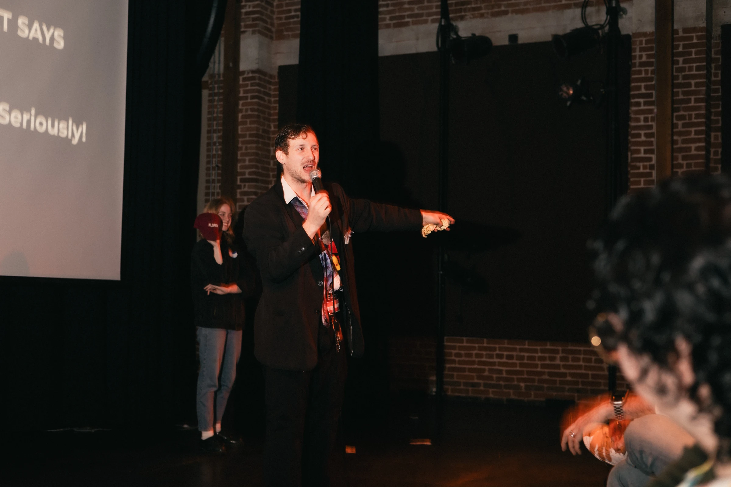 Man in a black suit holding a microphone points forward on a dimly lit stage with a visible projection screen; casual, engaging atmosphere.