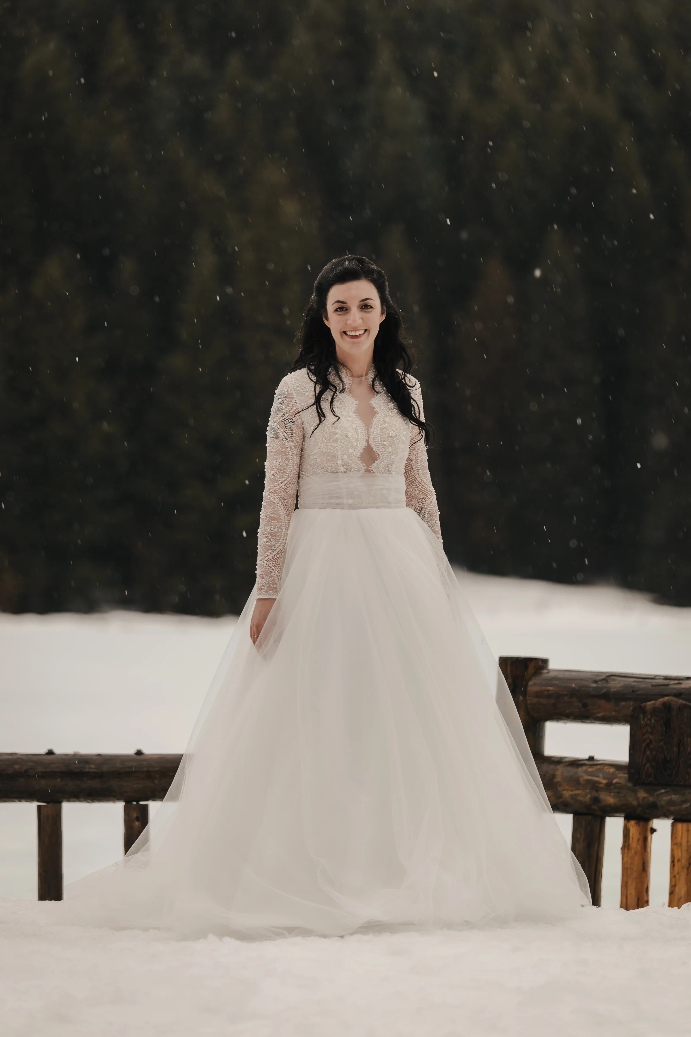 Bride in a white lace wedding dress stands in snowy landscape, smiling amidst falling snowflakes. Dark green forest provides a serene backdrop.
