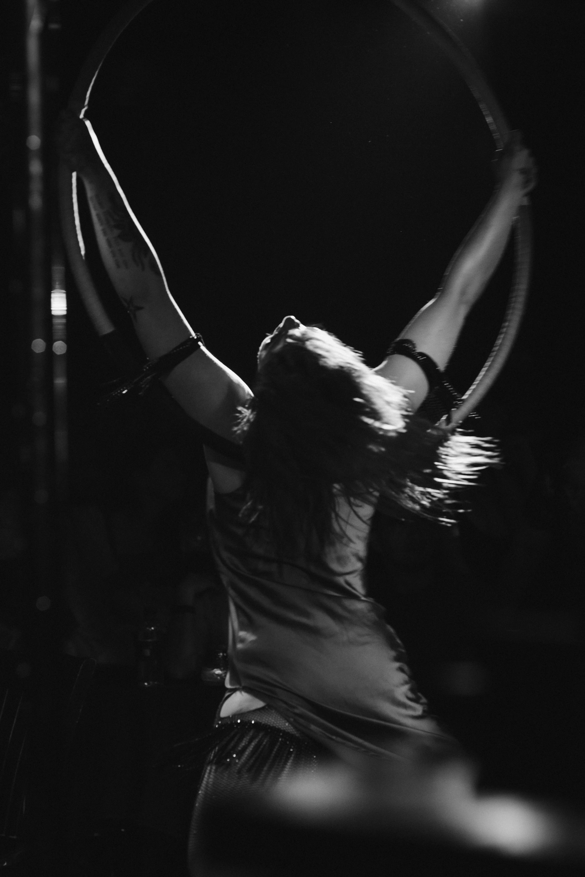 A performer is captured in mid-dance, silhouetted with dramatic lighting, holding a circular hoop overhead. The image conveys dynamic energy and focus. Black and white tones highlight the movement.