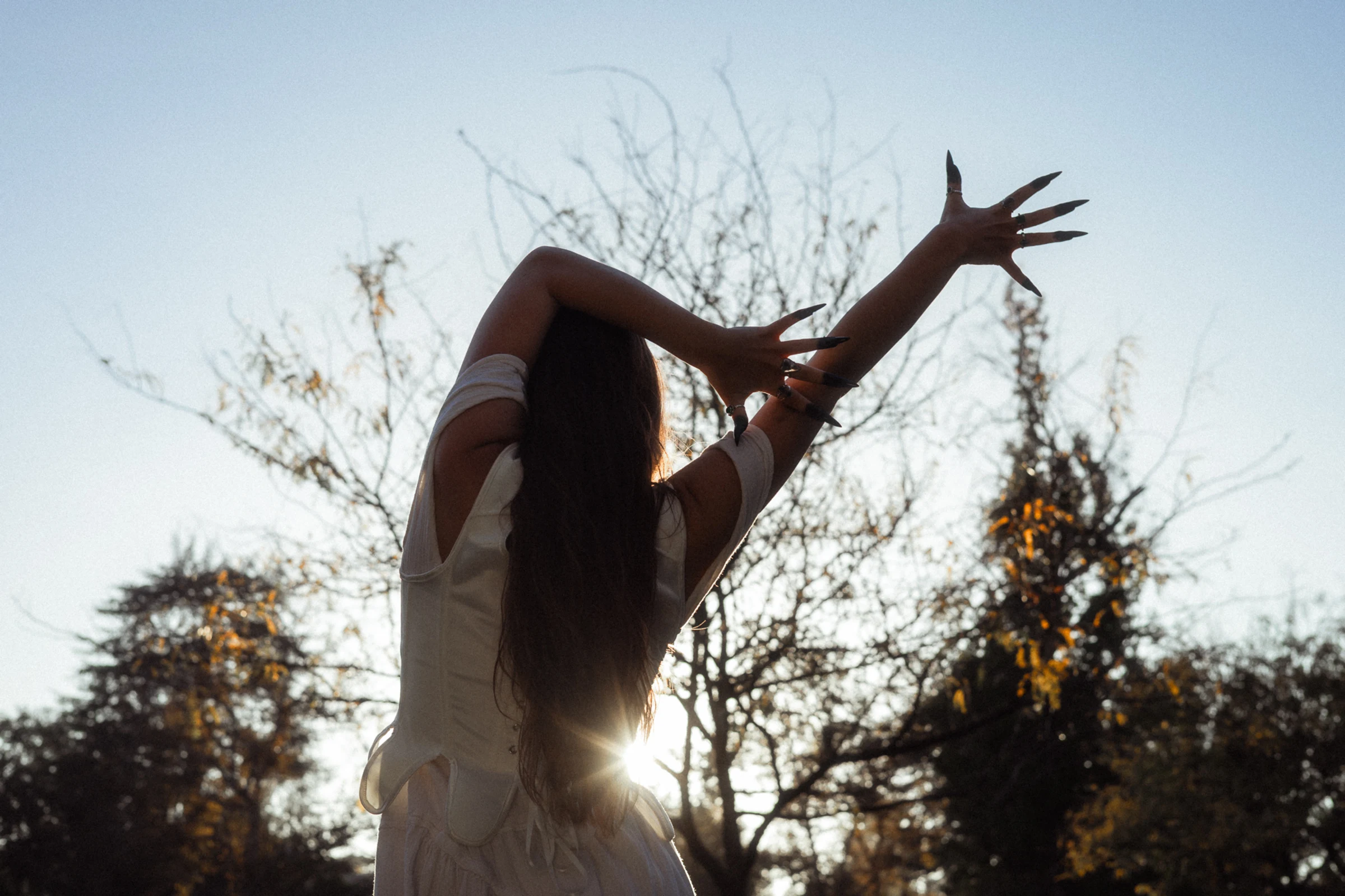 A person in a white dress gracefully poses with arms outstretched against a bright blue sky, silhouetted by sunlight peeking through bare trees, evoking serenity.