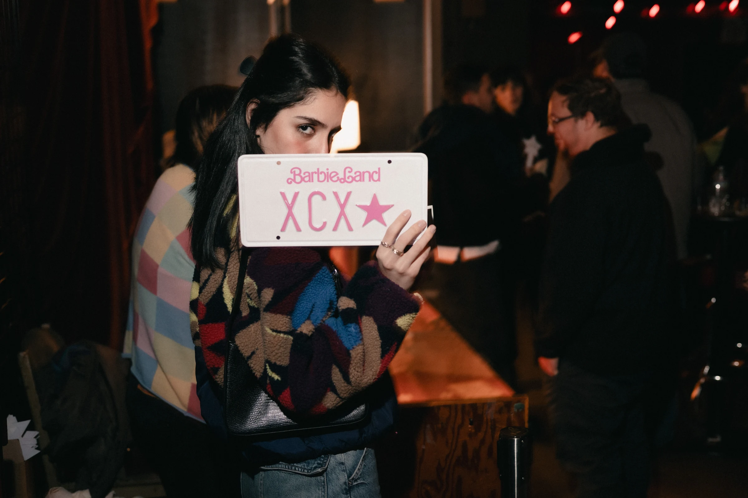 A person in a colorful sweater holds a "Barbie Land XCX" sign, looking at the camera. The setting is a dimly lit room with people socializing.
