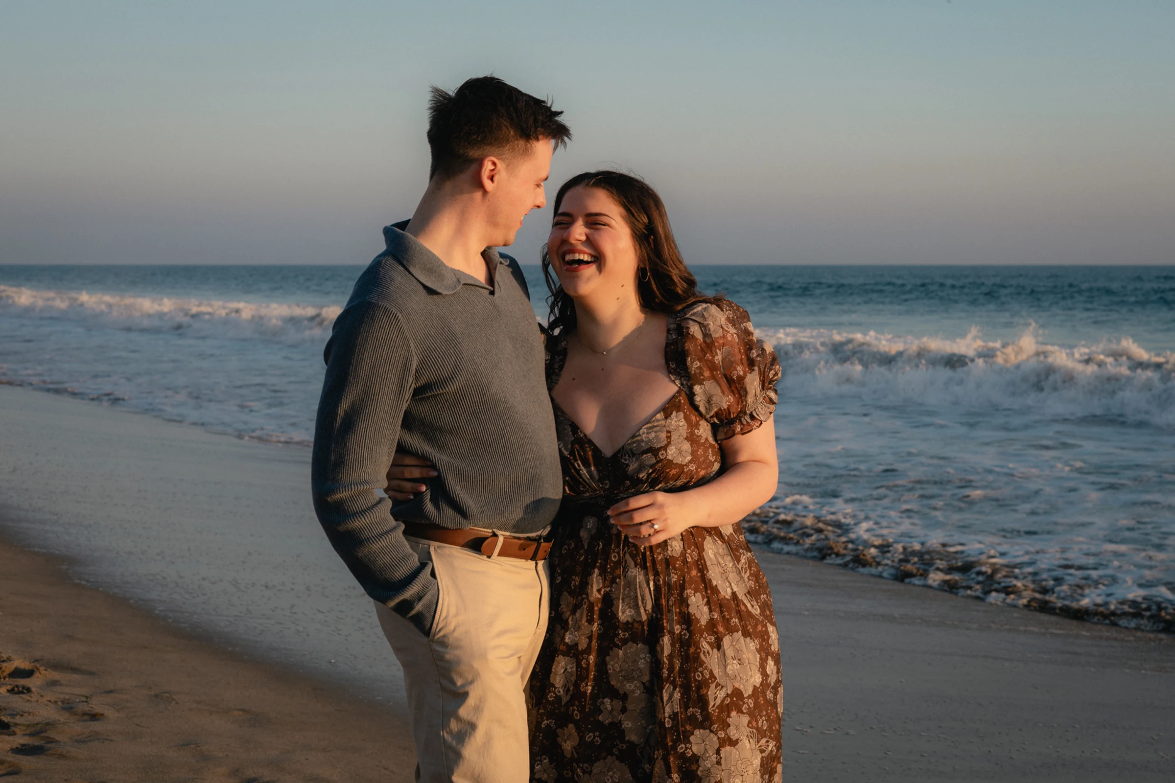 A couple laughs warmly on a beach at sunset. The woman wears a floral dress, and the man is in a blue sweater. Waves crash gently in the background.