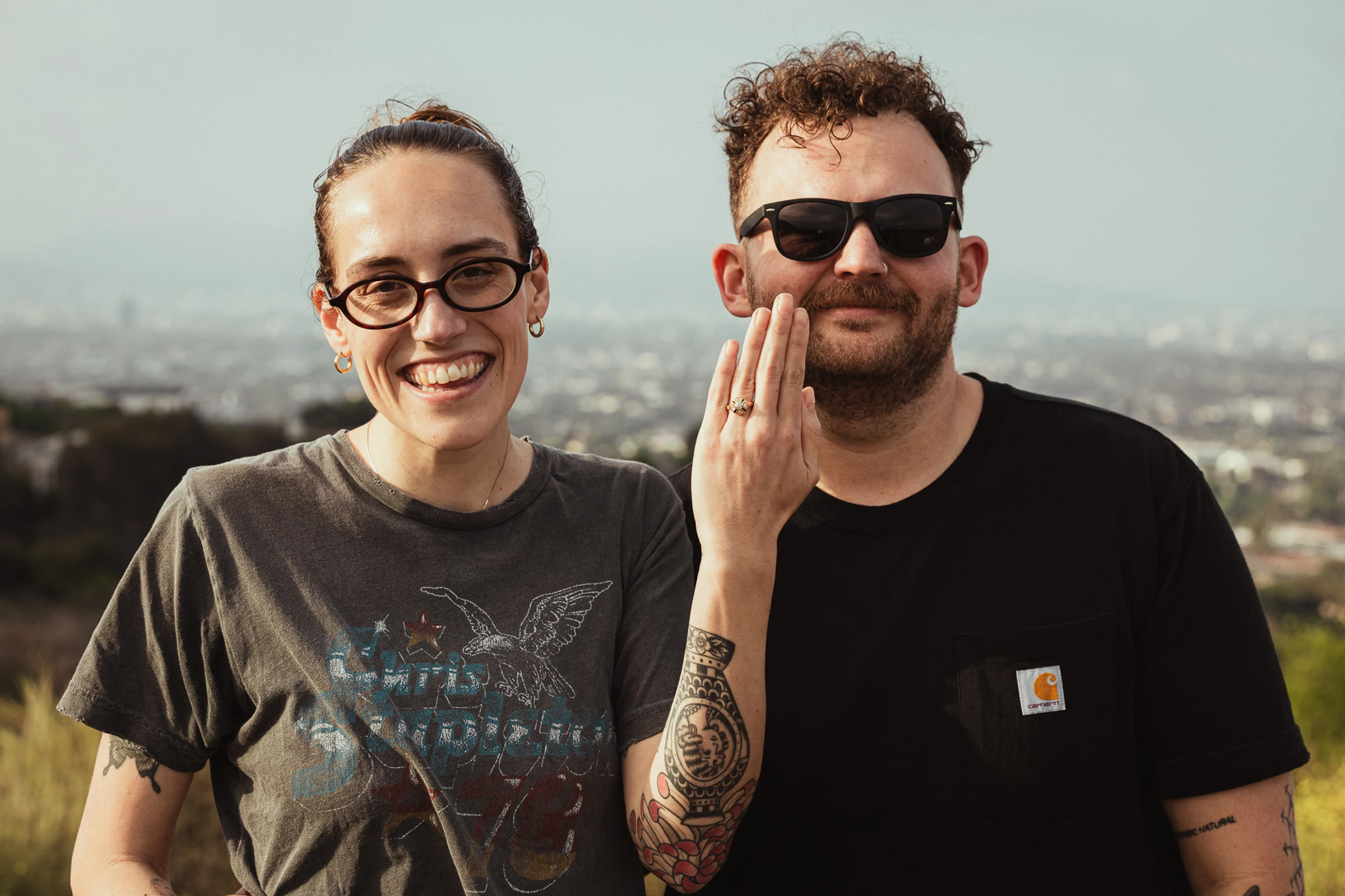 Two people stand outdoors smiling at the camera, one displaying a ring on their hand. A cityscape is visible in the blurred background.