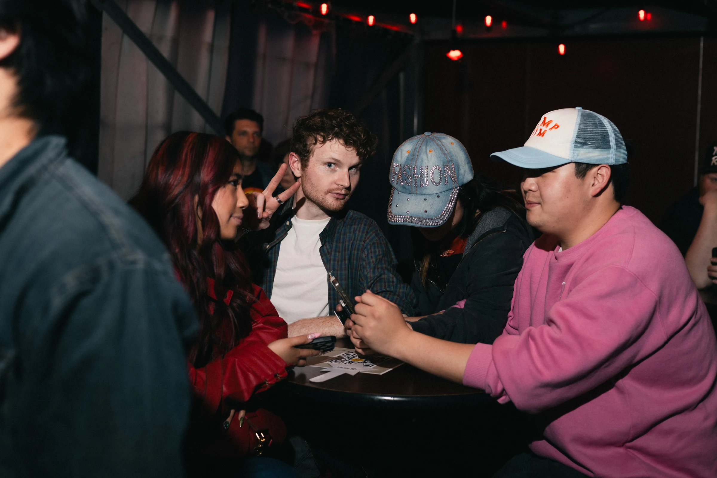 Group of people seated around a small round table at a dimly lit indoor venue, some wearing casual hats and one person making a peace sign.