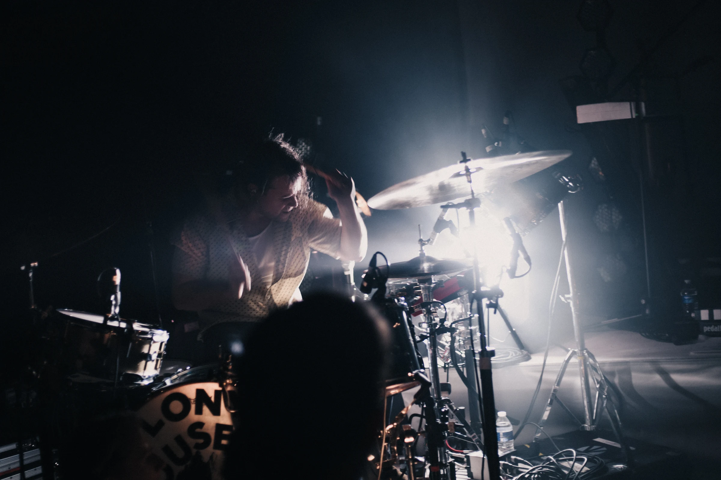 Drummer playing intensely in a dimly lit concert setting, illuminated by a bright spotlight. The scene conveys energy and focus, with dynamic shadows.