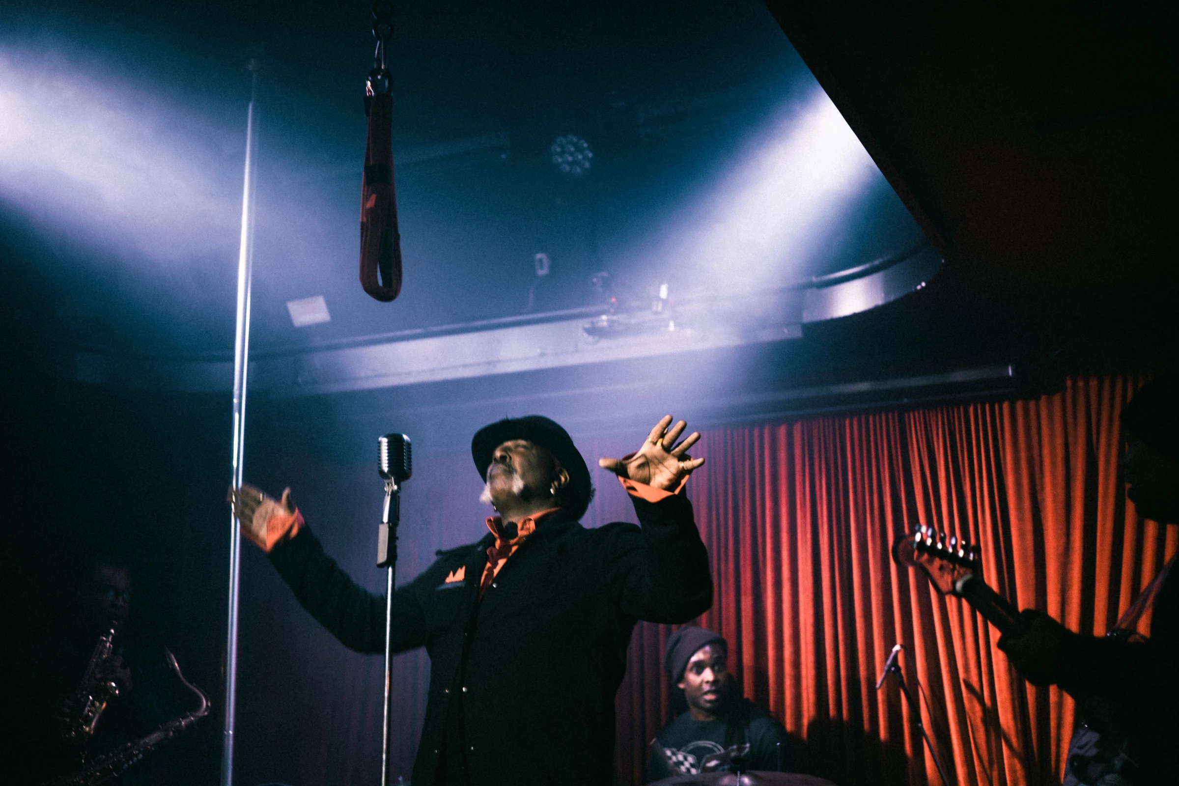 A musician in a dark suit and hat passionately sings into a vintage microphone on stage, surrounded by red curtains and band members, creating a moody jazz ambiance.