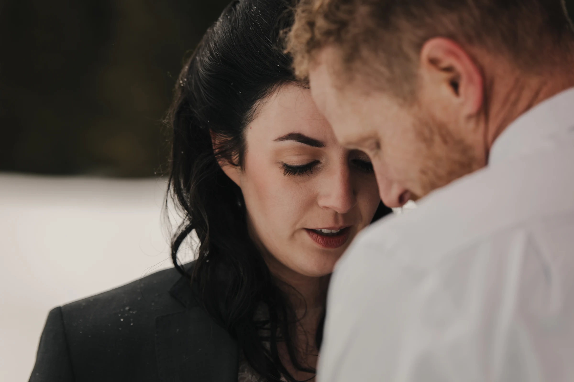 A couple, close together, shares an intimate moment in a snowy landscape. The woman looks down with a tender expression, and the man’s face is turned toward her.