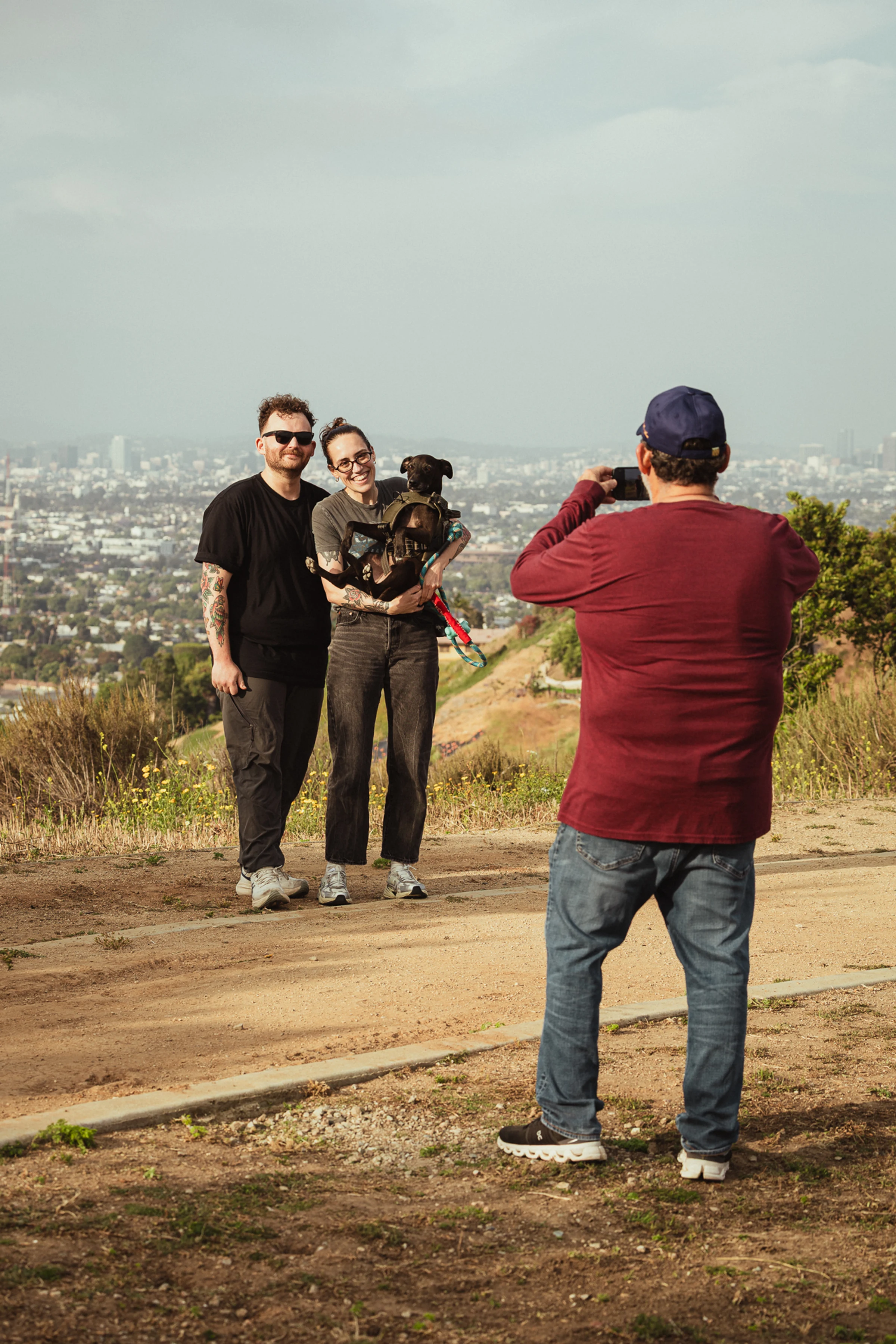 A person in a red shirt takes a photo of a smiling couple with a dog. They stand on a trail with a cityscape view in the background. The mood is cheerful.