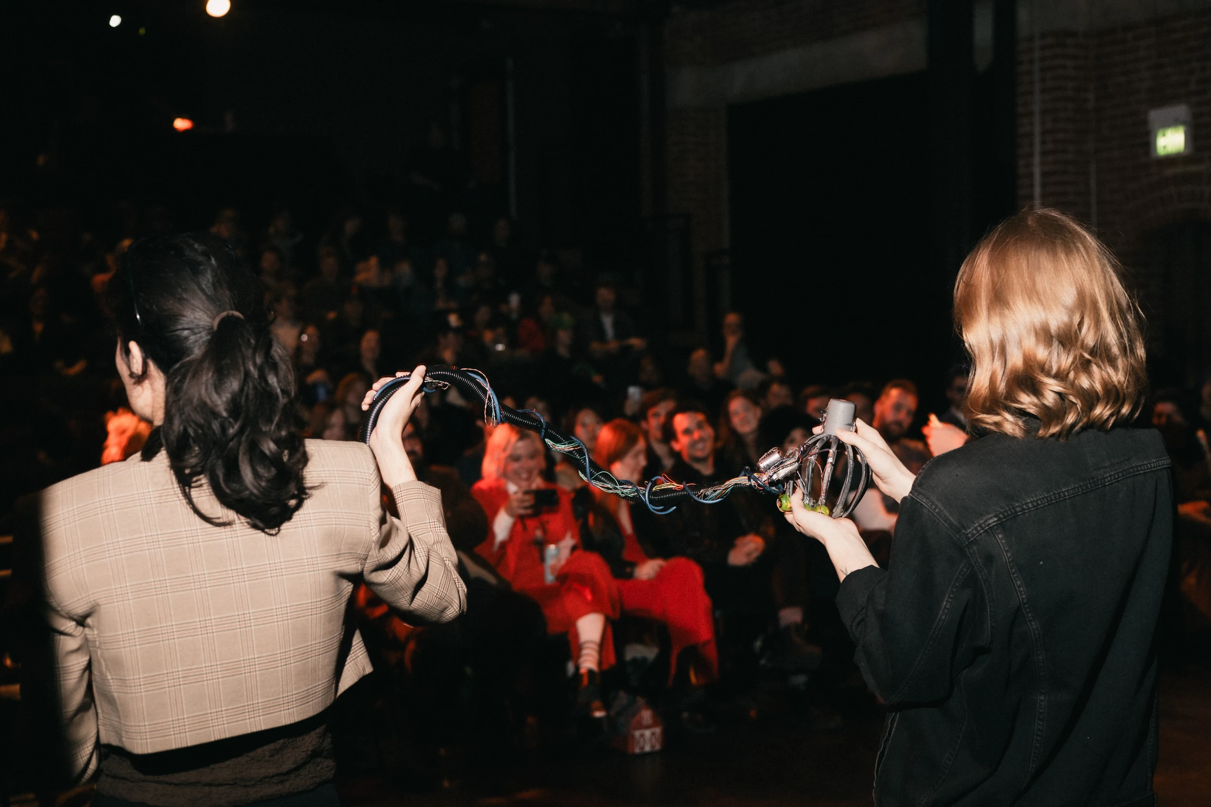 Two presenters on stage, facing an audience, holding a coiled cable or rope. The audience appears engaged and curious, with dim lighting creating an intimate atmosphere.