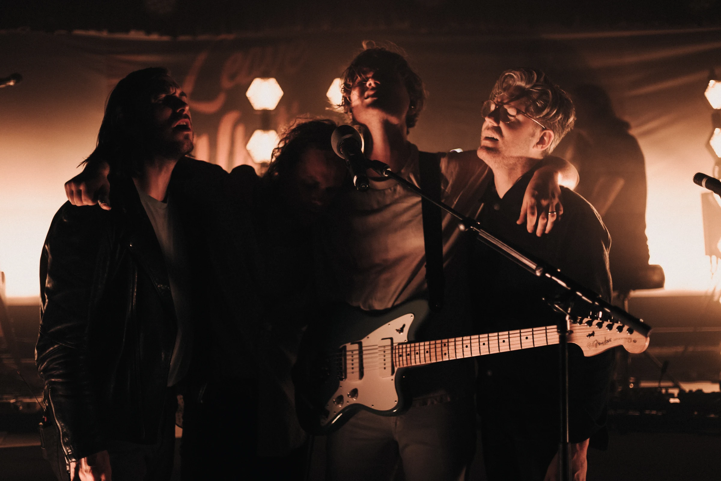 Four musicians stand close together on stage, lit by warm backlighting. They hold guitars, creating an intimate, soulful atmosphere.