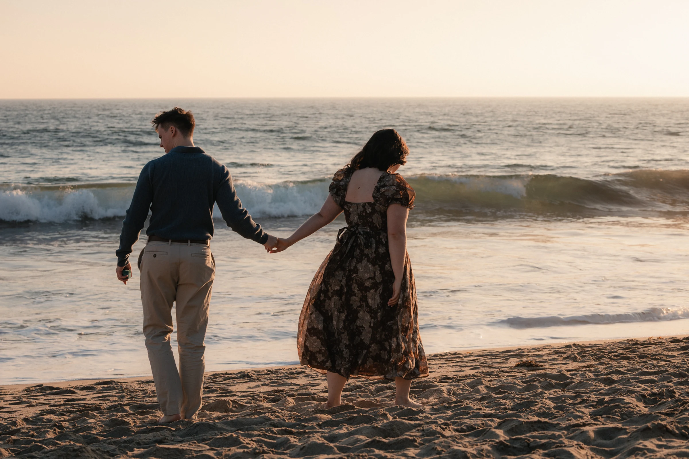 A couple holds hands while walking along a sandy beach at sunset. Waves gently crash in the background, creating a serene and romantic ambiance.