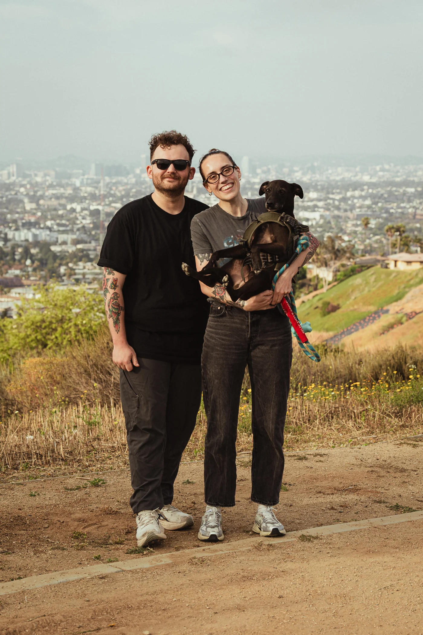 A smiling couple stands on a dirt path overlooking a cityscape. The man wears sunglasses and black clothes. The woman, in a gray t-shirt, holds a small black dog.