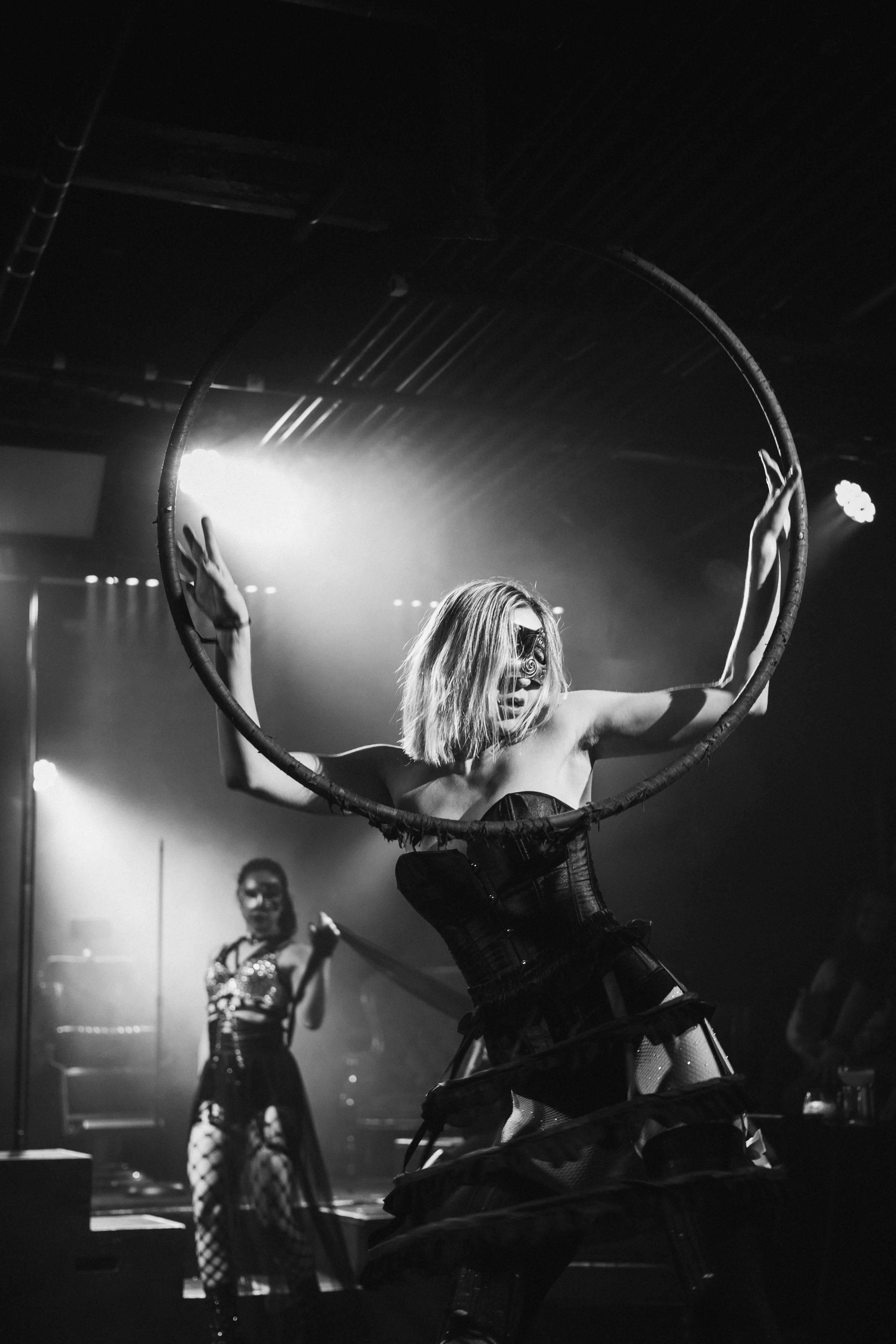 A performer in a corset poses dramatically with a large hoop on stage, backlit by spotlights. Another costumed figure stands in the background. Moody atmosphere.