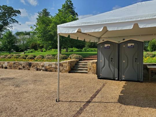 Two APT portable toilets positioned under a white event tent at a garden venue