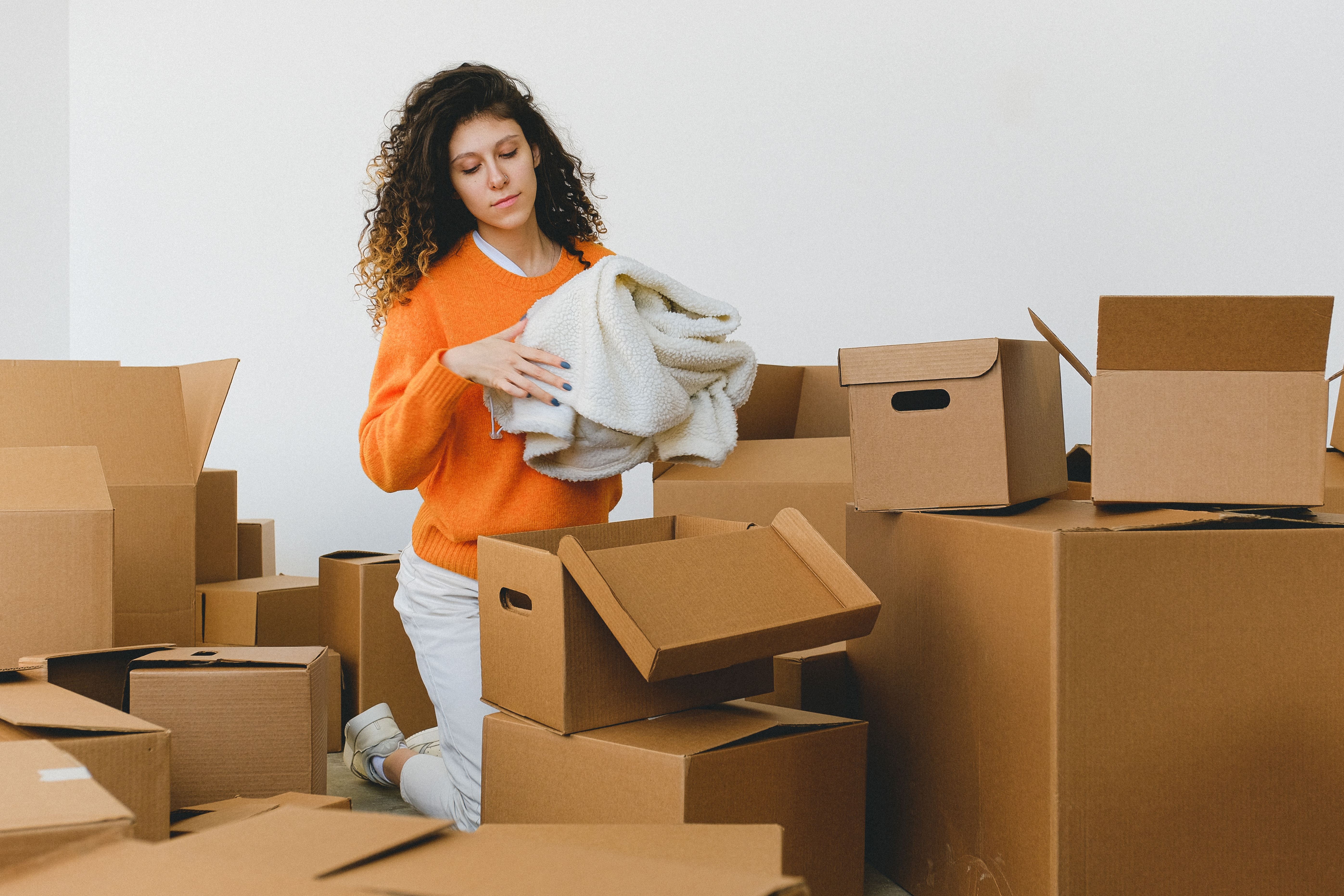 Image Woman in orange sweater kneeling among moving boxes, packing belongings during a home relocation