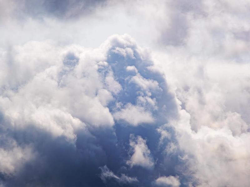 A detailed photograph of a dense, billowing cumulus cloud. Its tops are bright white where they catch the sunlight, while its base is a deep, shadowy blue-grey.