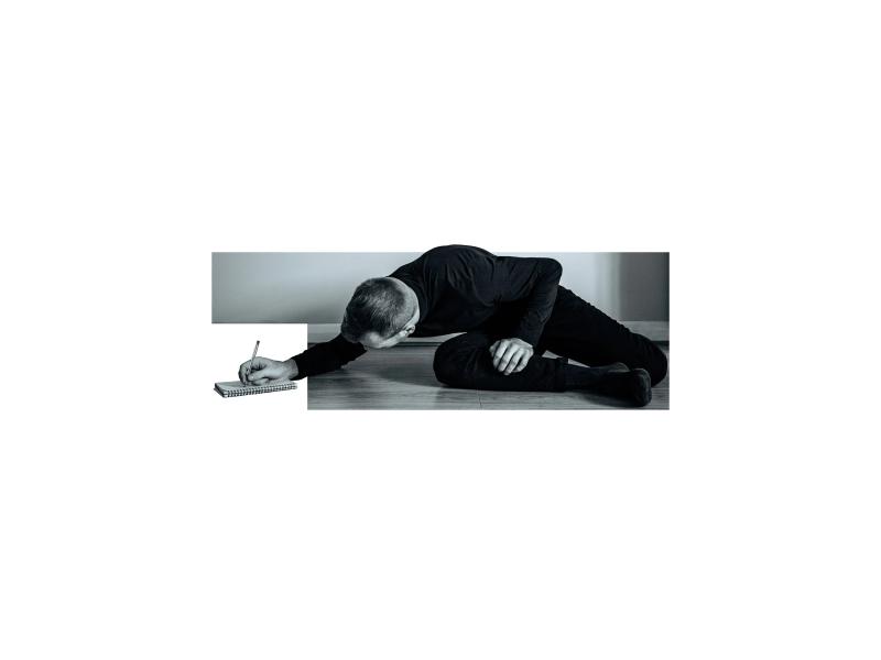 A conceptual black and white photo showing Dylan Burnside-Smith writing on the floor, with his hand and notebook extending beyond the image's tight border.