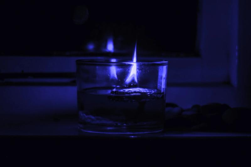 A moody, blue-toned photograph of a lit candle in a glass jar sitting on a windowsill at night. The flame is the only source of light, reflecting in the dark window behind it.