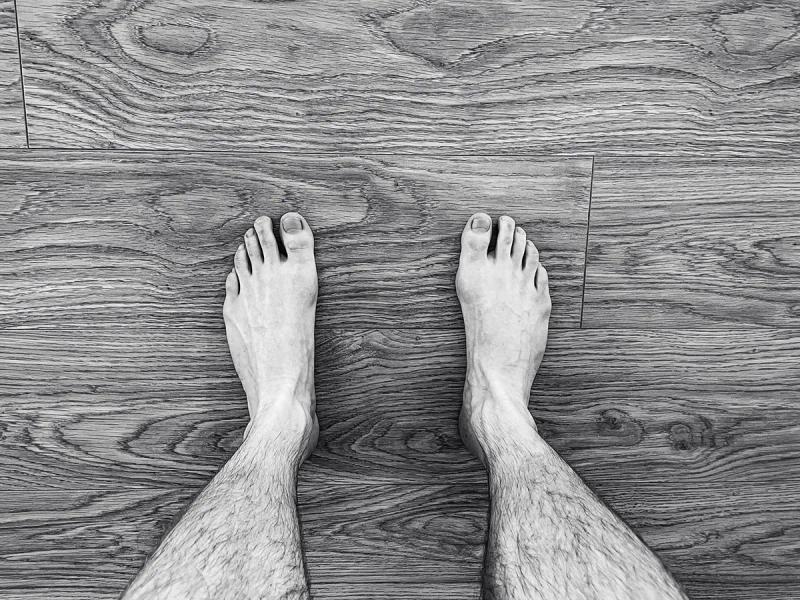 A first-person perspective in black and white of bare feet standing on a wooden floor, symbolizing the concept of taking the first step.