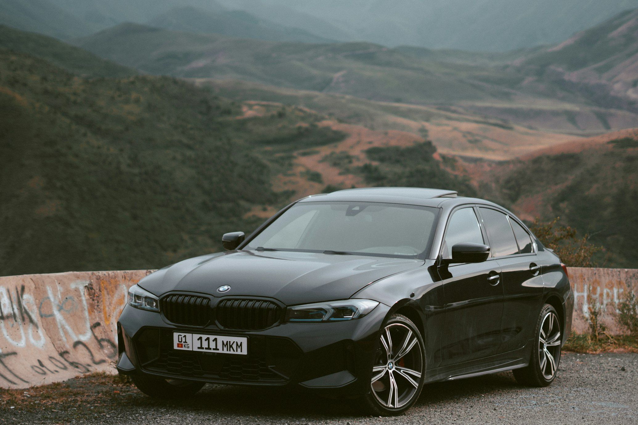 Black BMW 3 Series saloon parked beside a mountain road, viewed from the front three-quarter angle.