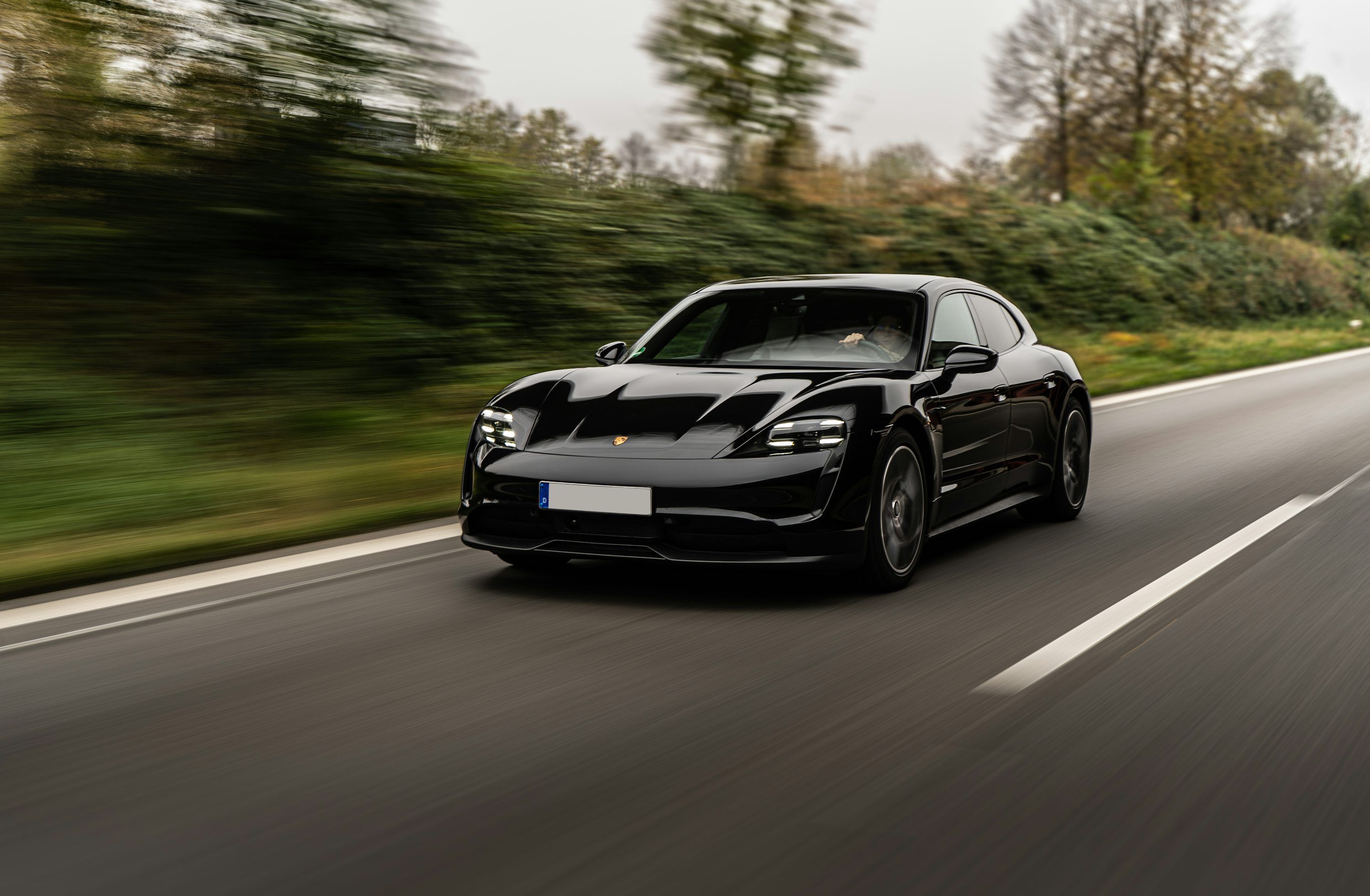 Black Porsche Taycan electric saloon travelling along a tree-lined road.