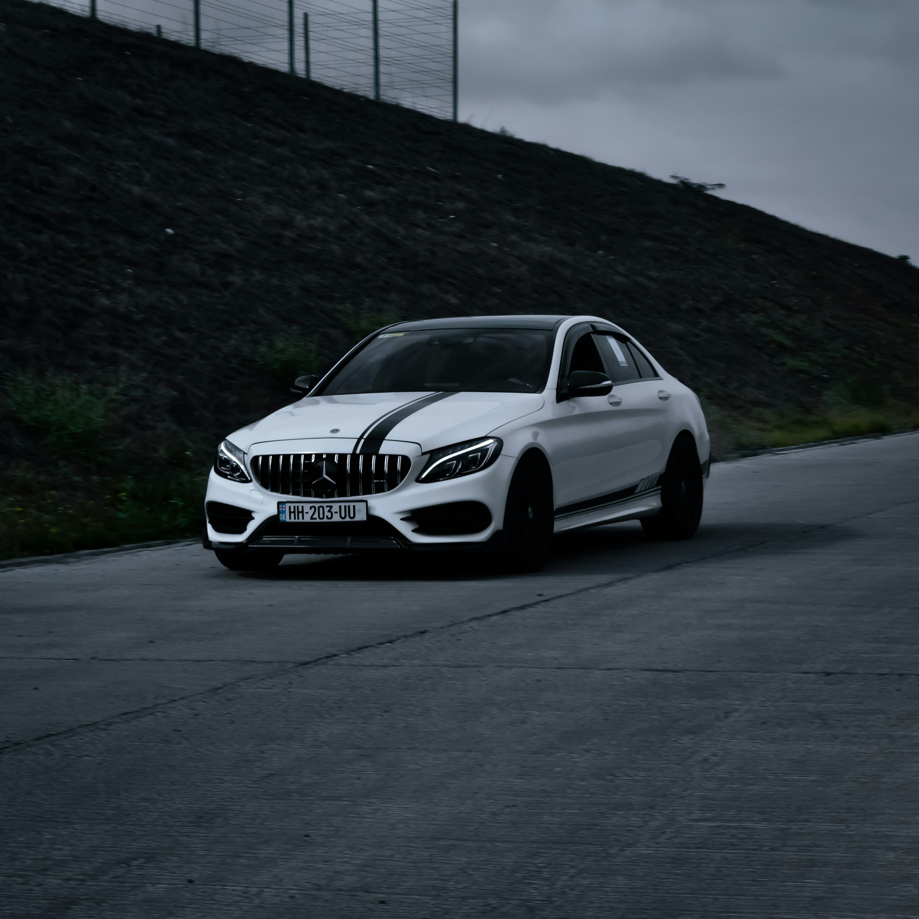 White Mercedes-Benz C-Class saloon with black bonnet stripes on a road beside a stone embankment.