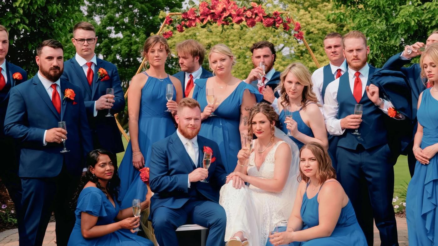 Large group in blue, black, and pink formal clothing posed outdoors beside red flowers.