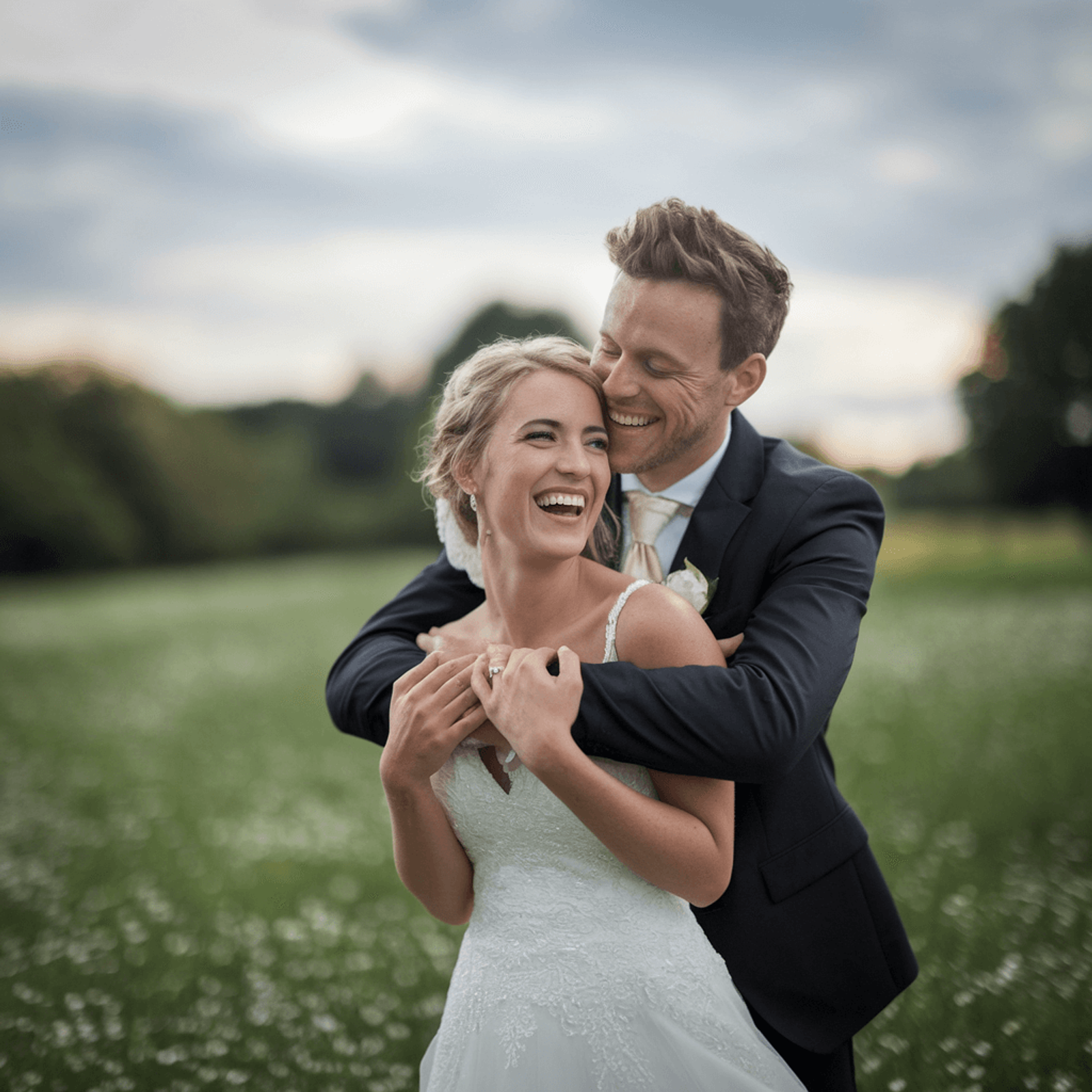 Couple in formal clothing embracing in a grassy field beneath a cloudy sky.