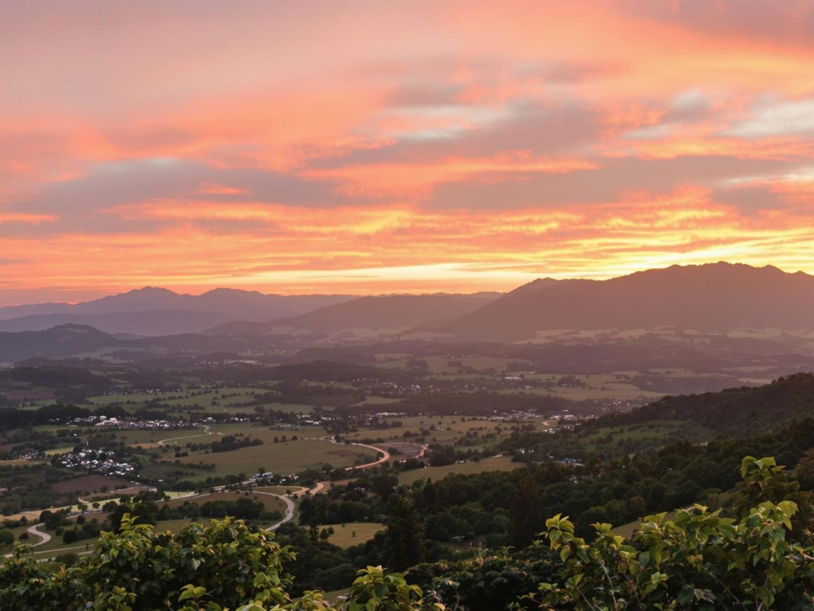 Orange and pink sunset above a valley, fields, and distant hills.