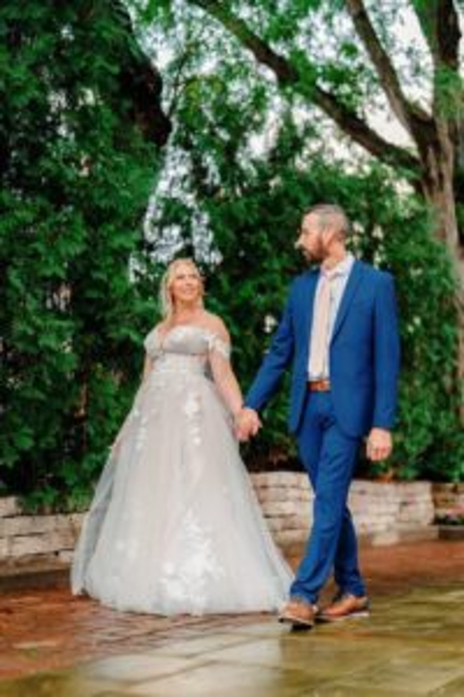 Couple in formal clothing walking together beneath a large leafy tree.