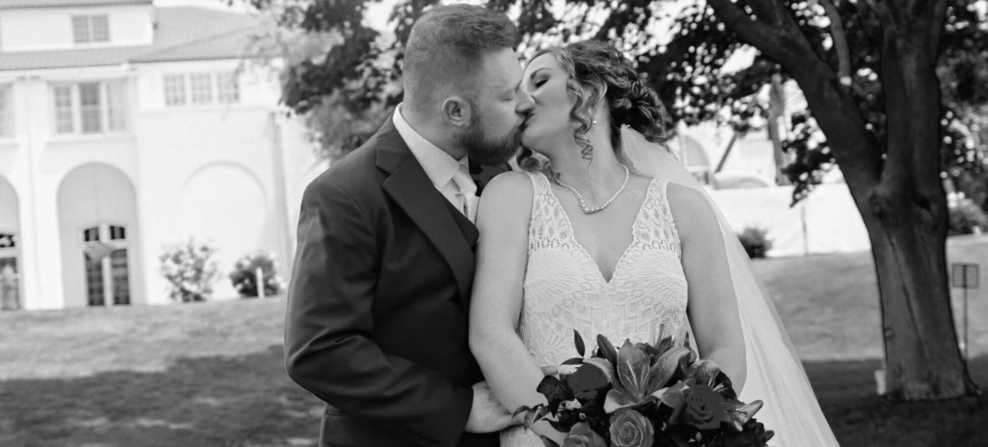 Black-and-white portrait of two formally dressed people kissing beneath trees near a white building.