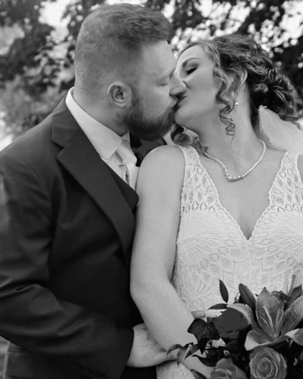 Tree Shade Kiss Black-and-white portrait of two formally dressed people kissing beneath trees near a white building.