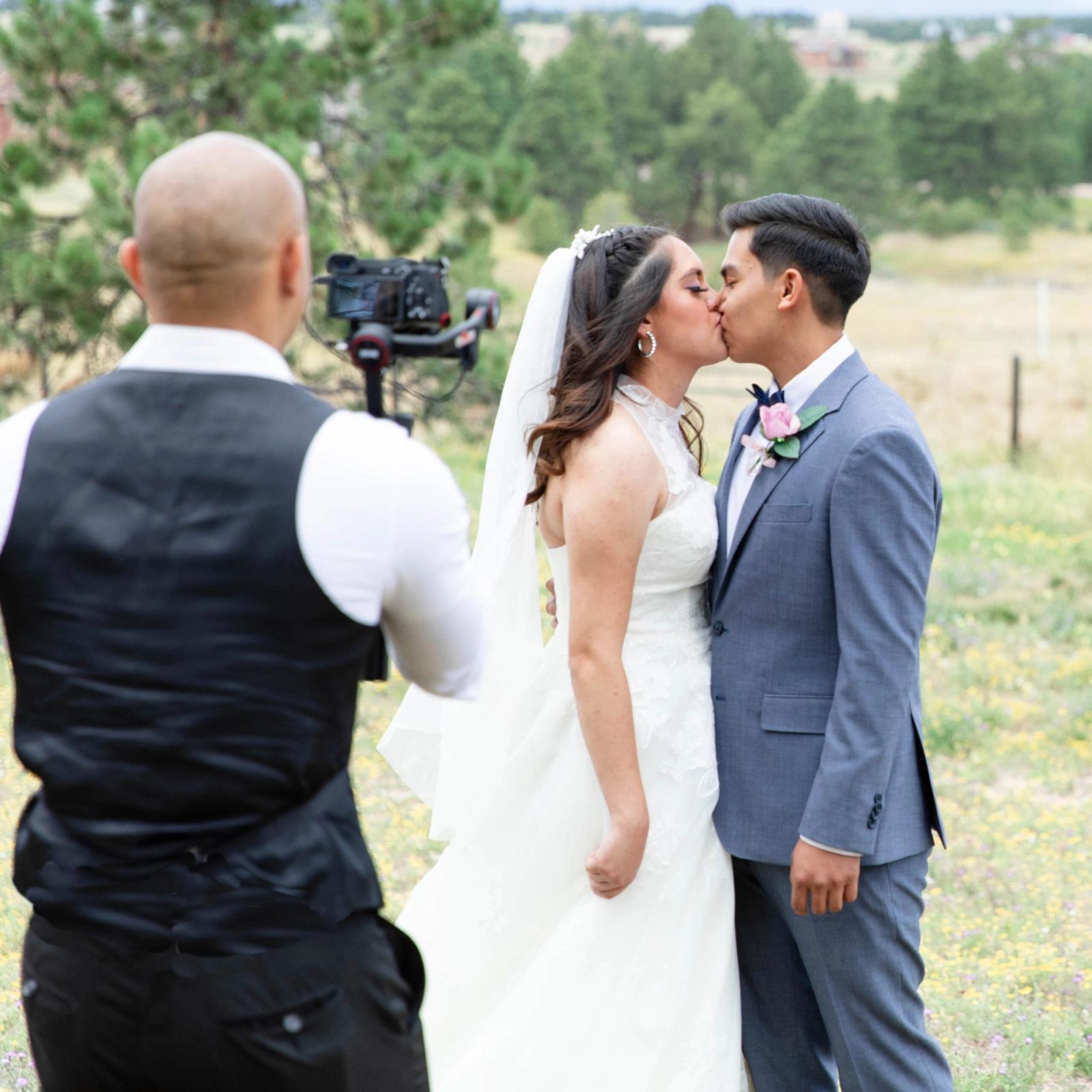 Camera operator films a couple kissing outdoors while another person stands nearby.