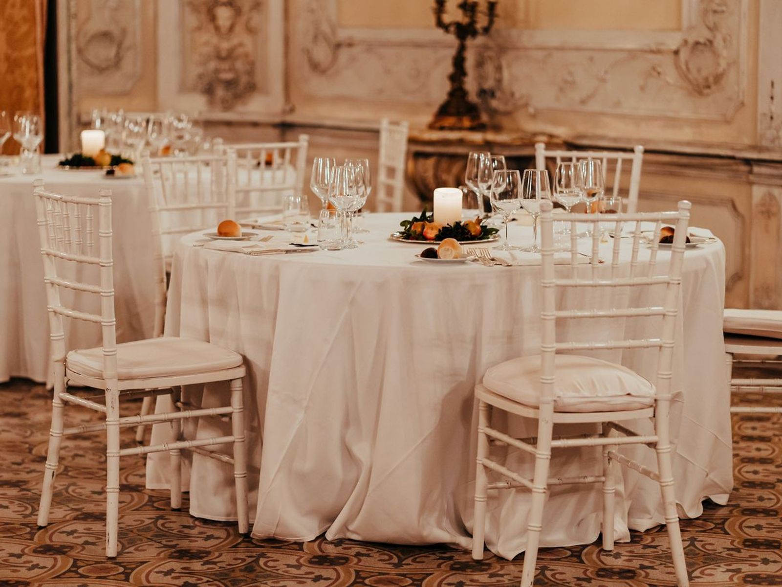 Round dining tables set with white linens, glassware, and pale chairs in an ornate room.