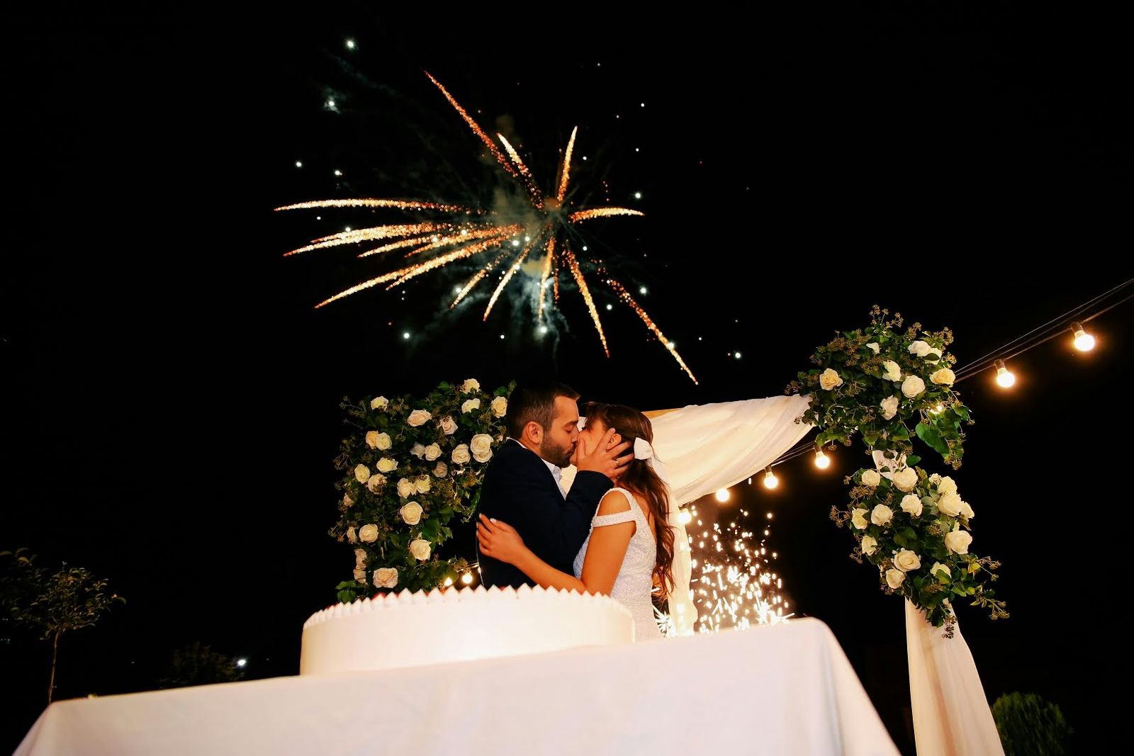 Fireworks Kiss Couple kissing behind a white table as fireworks and string lights glow overhead.