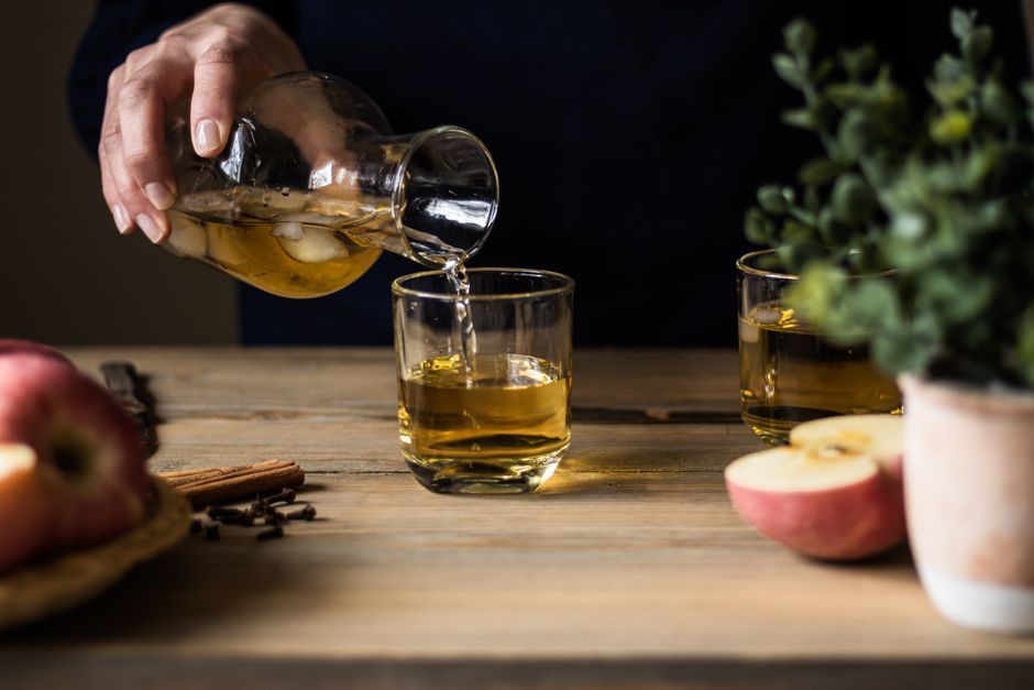 Cider Pour Hand pours amber liquid into a glass beside apples and cinnamon sticks.