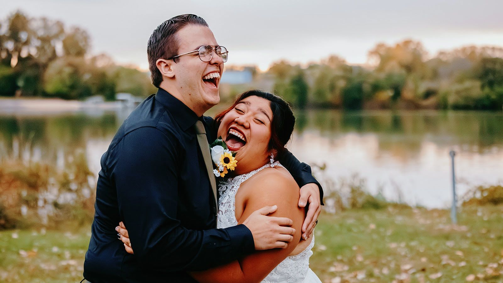 Couple in formal clothing laughing and embracing beside a lake.