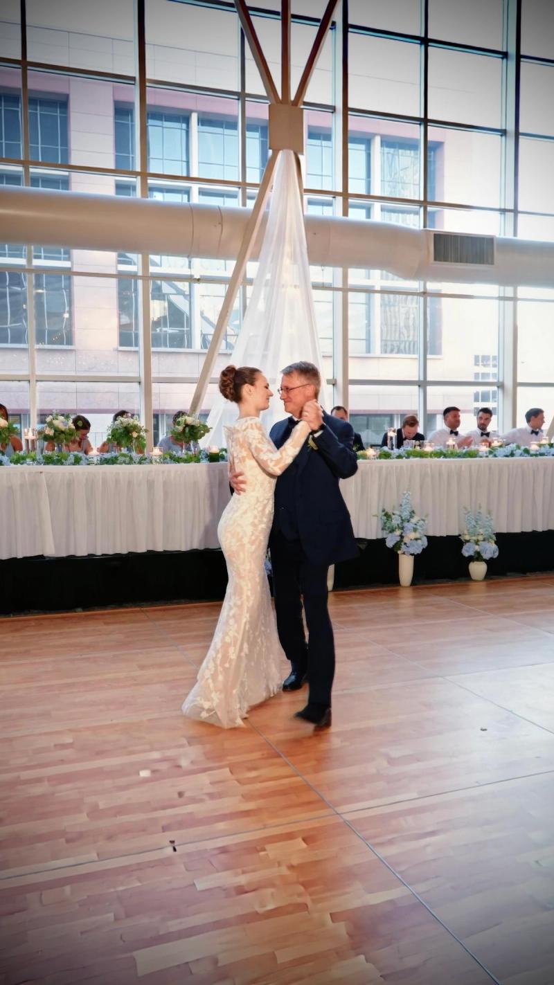 Two formally dressed people dancing in a bright hall with a long table behind.