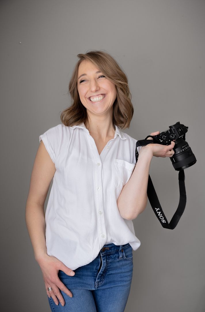 Photographer Portrait Woman in a white shirt holding a camera and smiling at the camera.