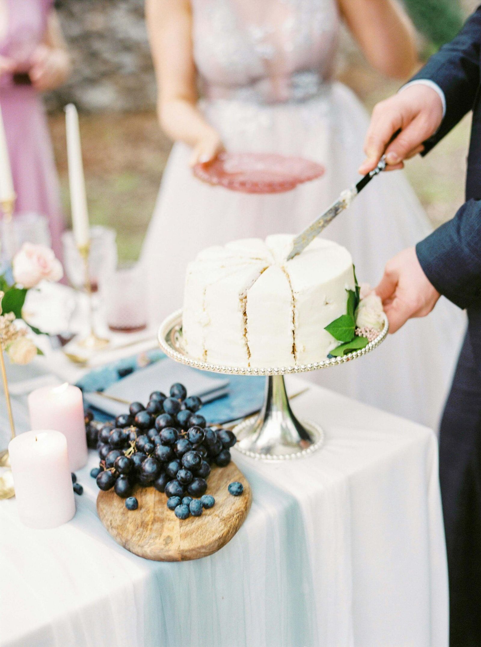 Hands cutting a white cake beside a bowl of dark berries.