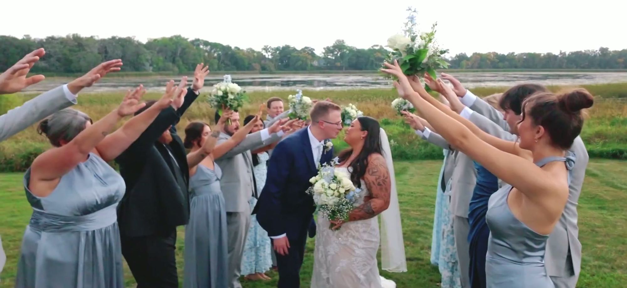 Couple kisses by a lake as guests raise their bouquets.