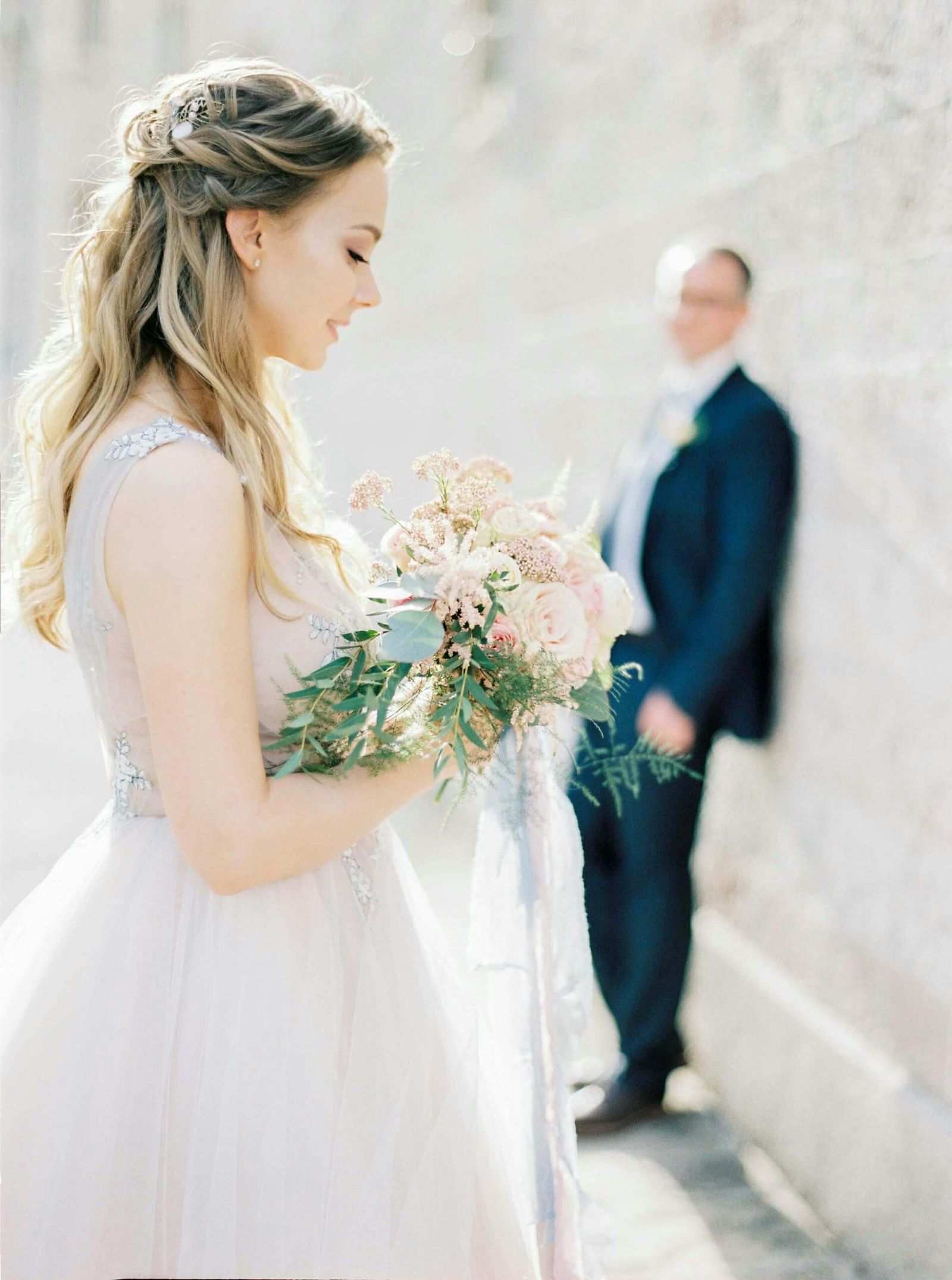 Person in a white formal dress holding a pale bouquet while another person stands blurred behind.