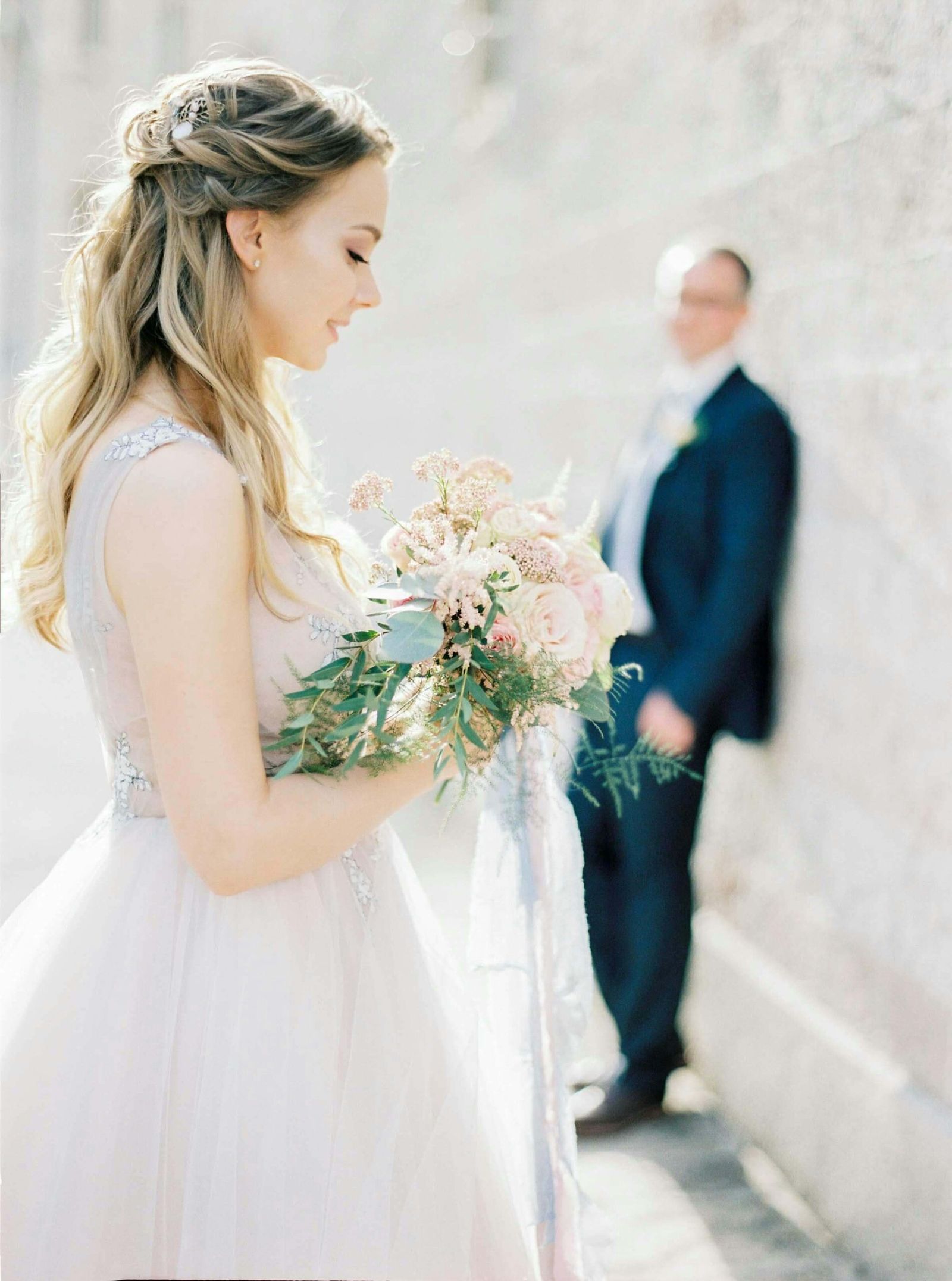 Pale Bouquet Portrait Person in a white formal dress holding a pale bouquet while another person stands blurred behind.
