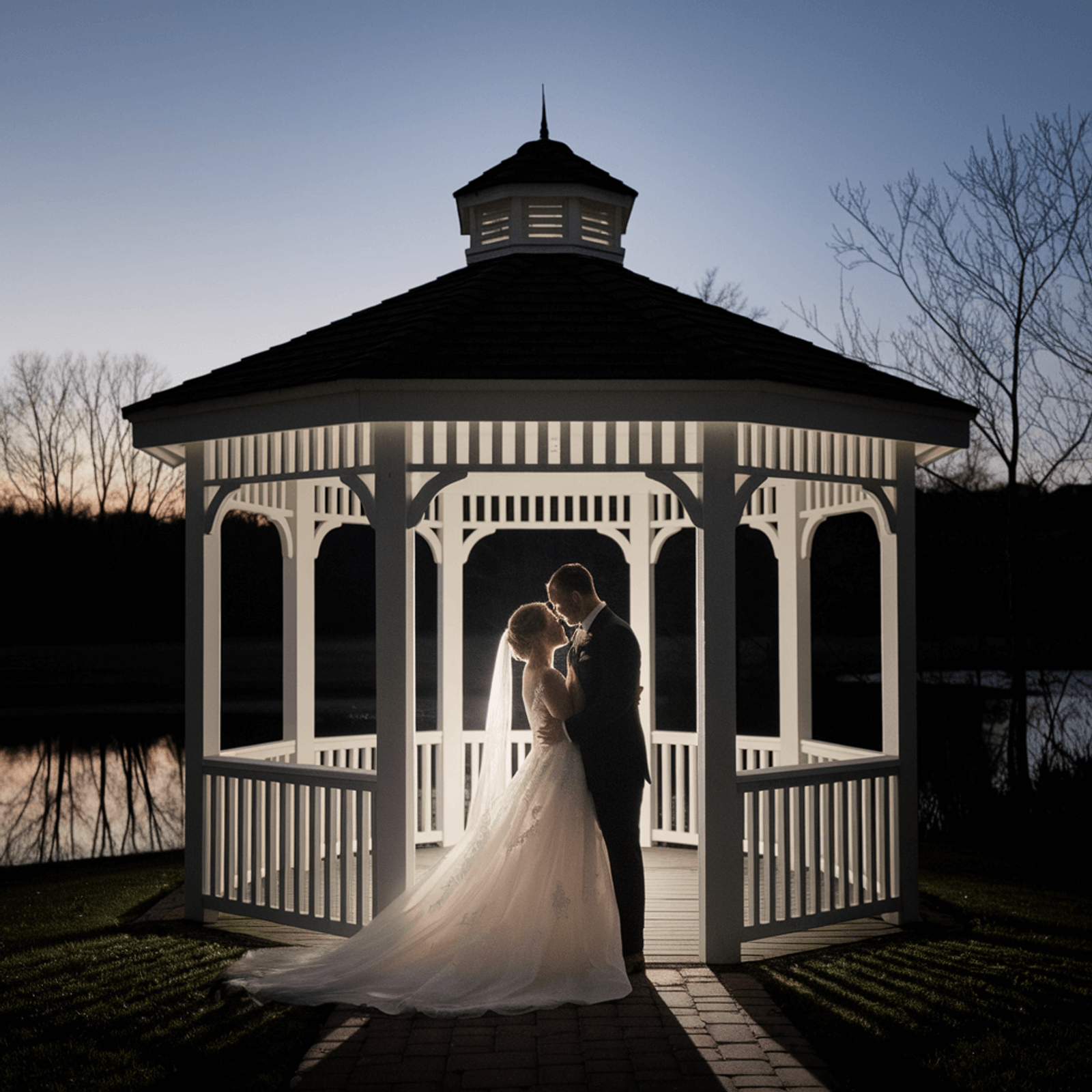 Couple in wedding attire embracing inside a white gazebo at dusk.