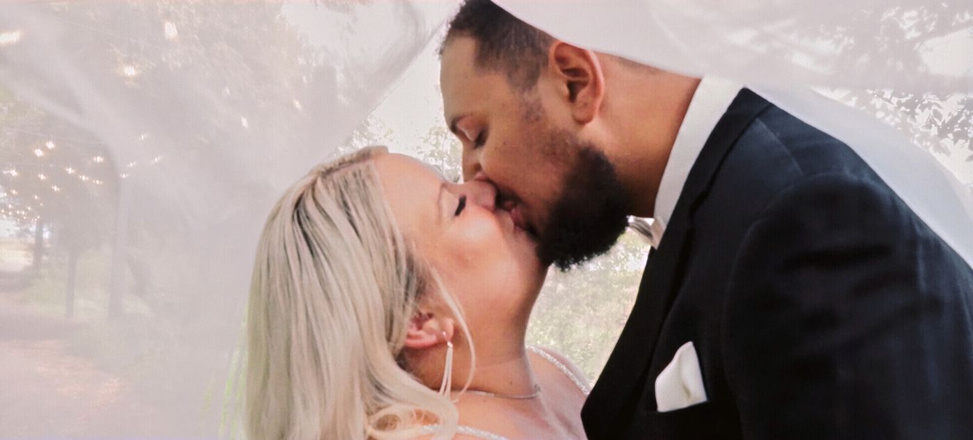 Close portrait of two formally dressed people kissing beneath translucent fabric.